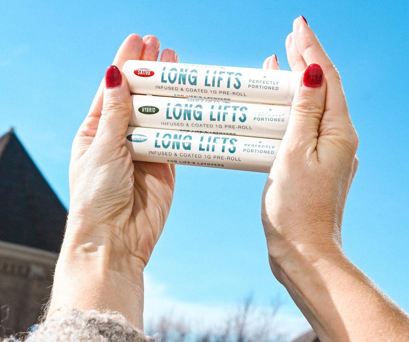 Three Long Lifts cannabis pre-roll packages labeled Sativa, Hybrid, and Indica held up against a clear blue sky.