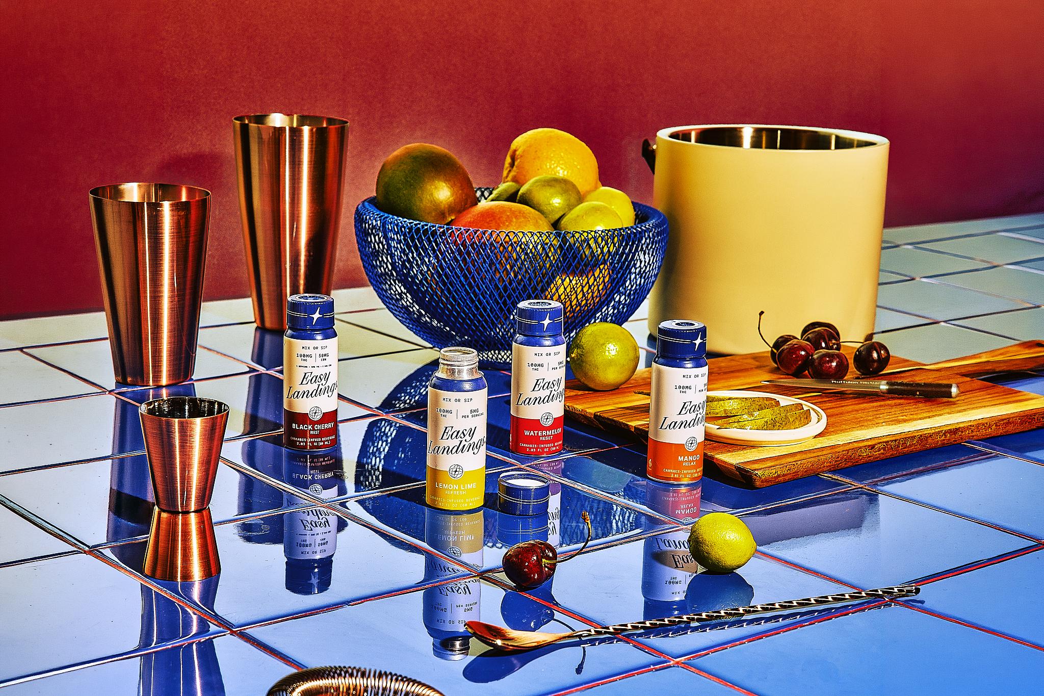 Easy Landing beverage products displayed with cocktail tools, citrus fruit, and cherries on a blue tiled surface.